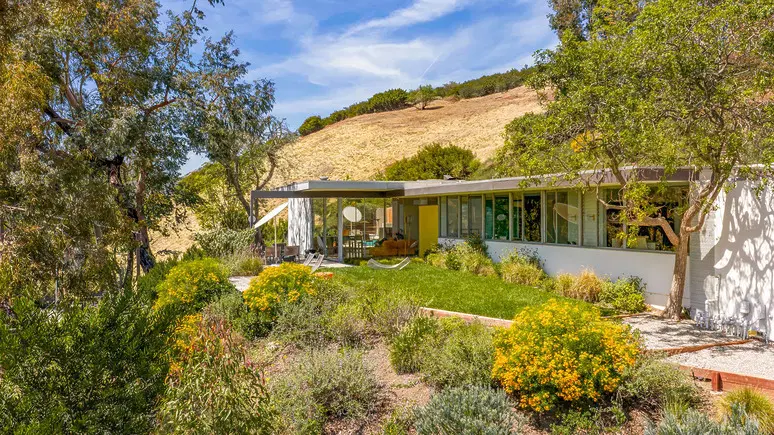 Mid-century modern house with large windows, surrounded by trees and hills under a clear blue sky.