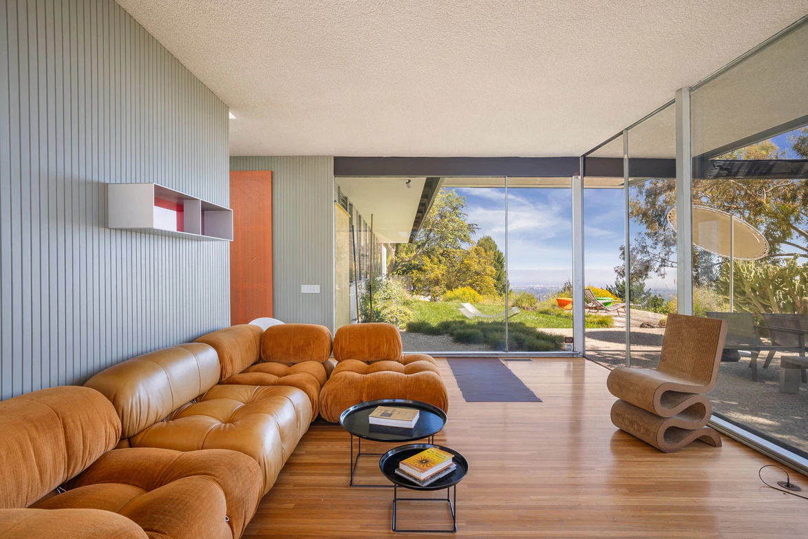 Modern living room with a brown leather sofa, glass walls, wooden floor, and a scenic outdoor view.