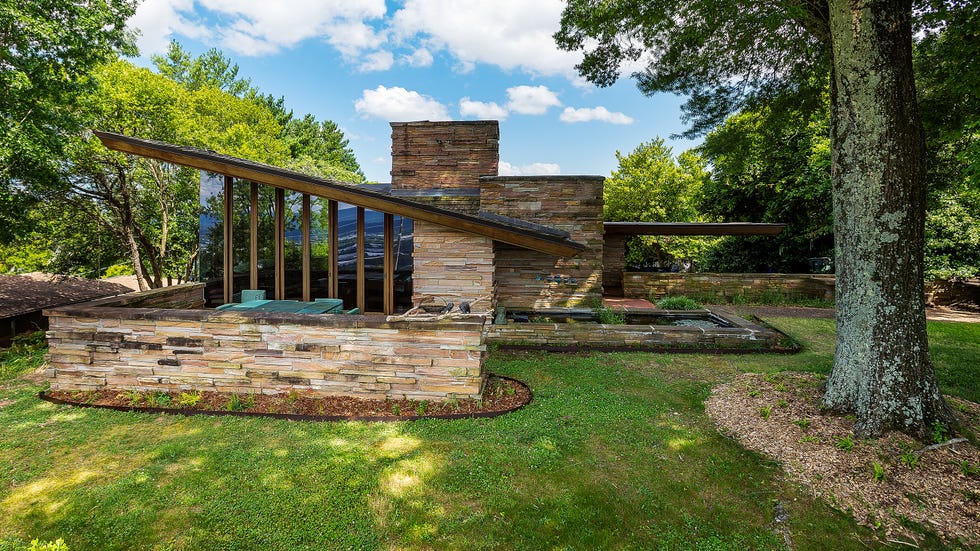 Modern stone house surrounded by greenery, featuring large windows and a unique roof design against a blue sky.