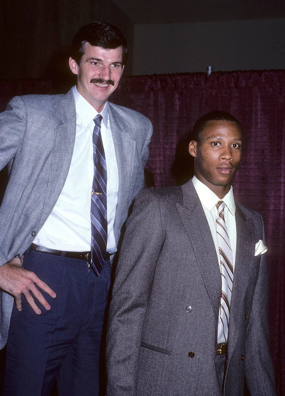 Two men in suits pose together at an event. One is tall with a mustache, the other has a serious expression. The setting suggests a sports gathering