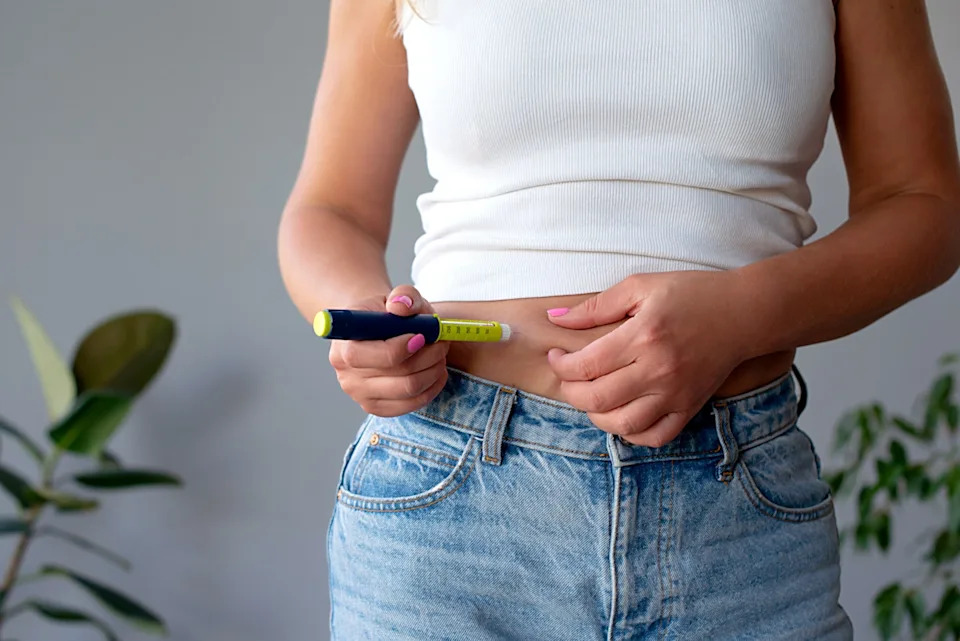 Person using an insulin pen on their abdomen, wearing a white top and blue jeans, symbolizing diabetes management