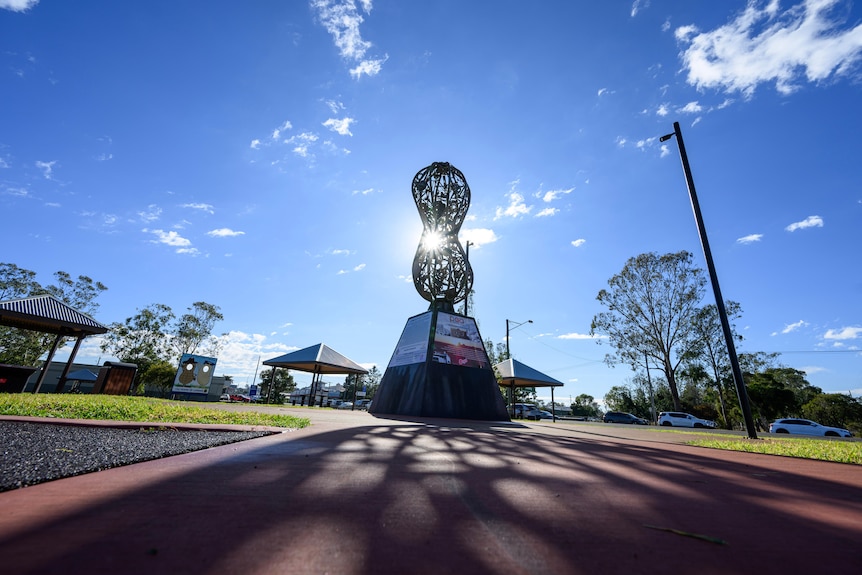 The sun shines through a giant peanut sculpture atop a stone stand in the middle of a park.