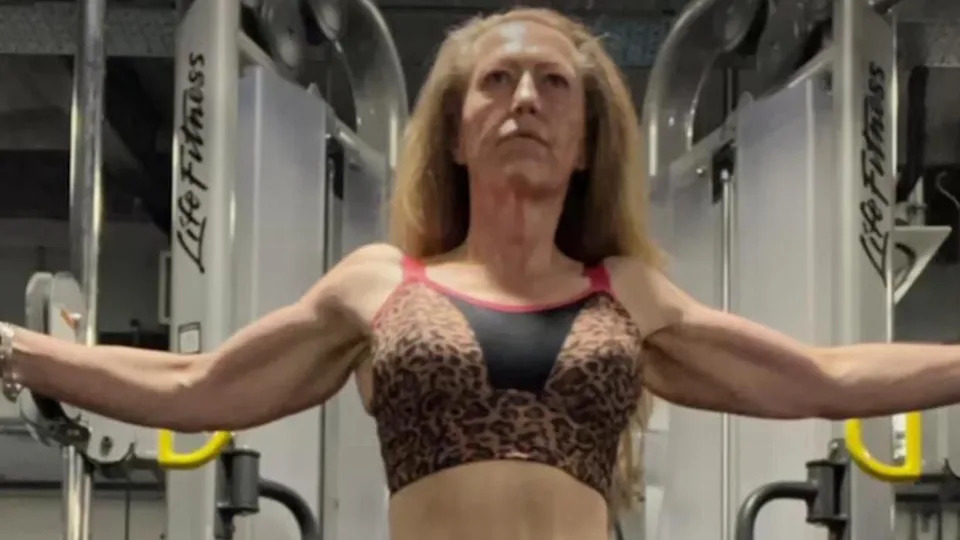 Jane Bennett training at the gym. She is stood flexing her muscles at a workout machine.