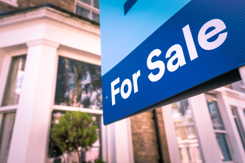 A blue "For Sale" sign for an estate agent, with white text, hanging in front of houses.