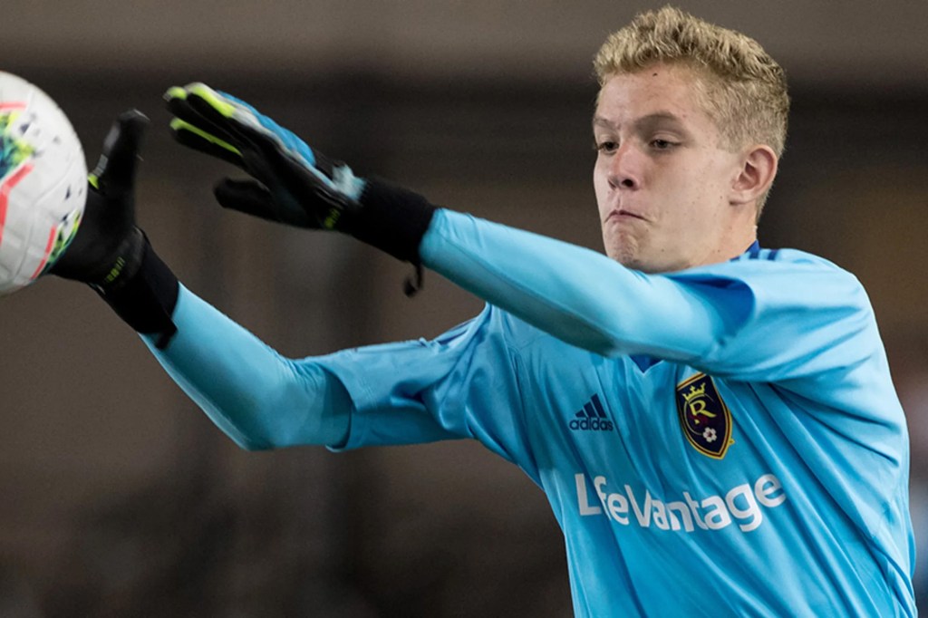 A male soccer goalie in a light blue jersey and black gloves catching a white and colorful soccer ball.
