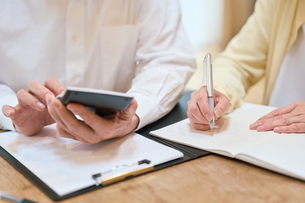 A person on the left holding a calculator, and a person on the right writing on a notebook.