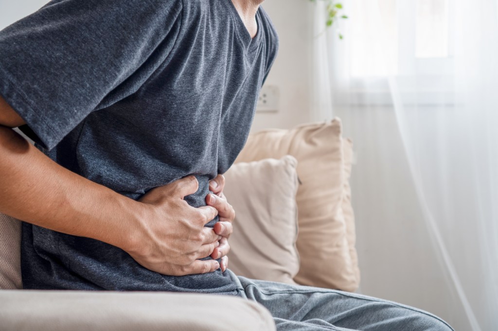 An Asian man sitting on a sofa, holding his stomach in pain.