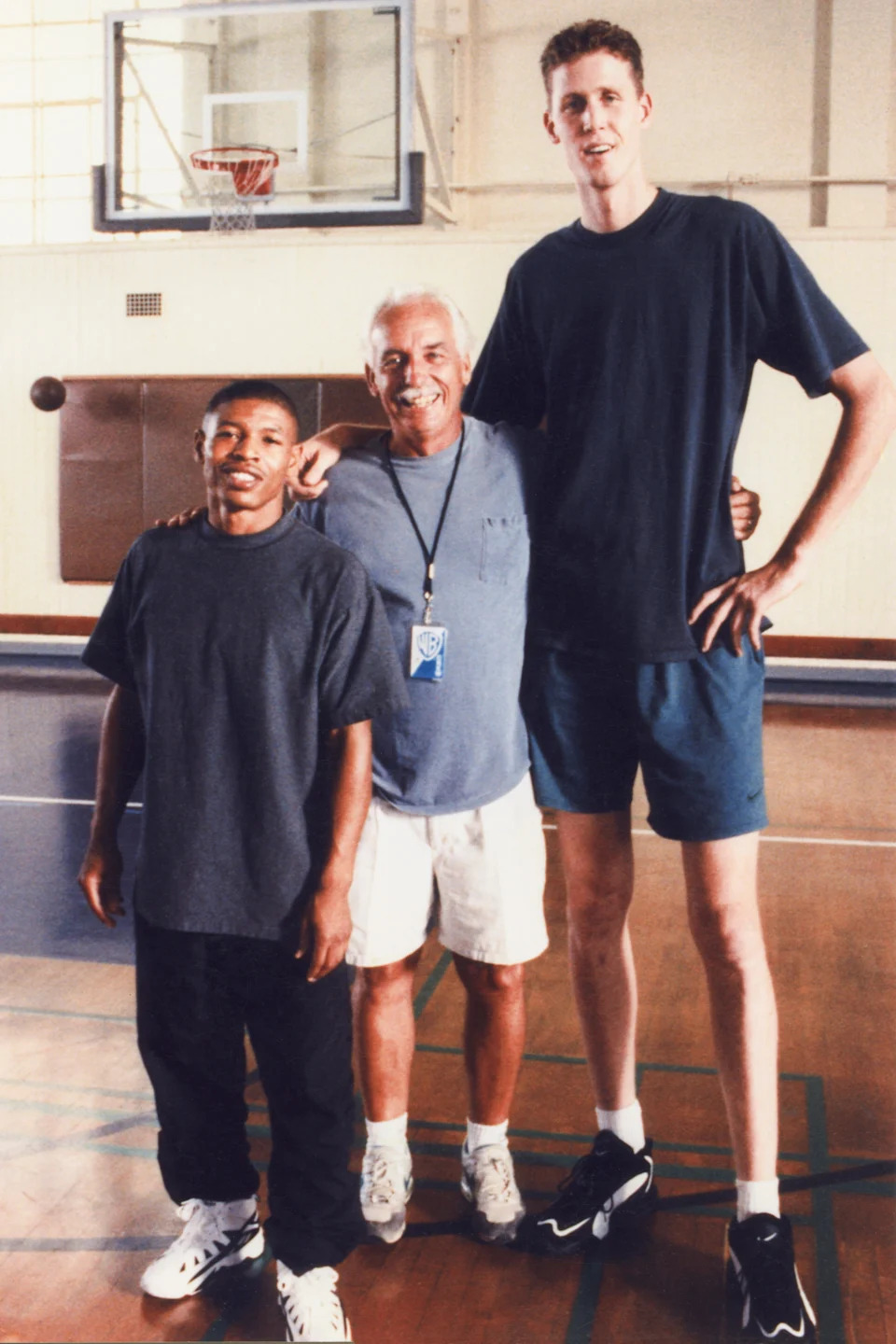 Three men standing in a gym, with one particularly tall man on the right. They all smile, suggesting a lighthearted atmosphere