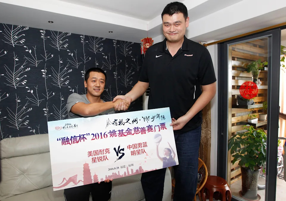 Two men shake hands while holding a large sign for a 2016 basketball charity event between U.S. and Chinese teams