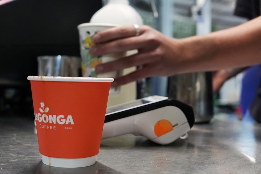 An orange paper coffee cup on a bench next to an espresso machine and a point of service.