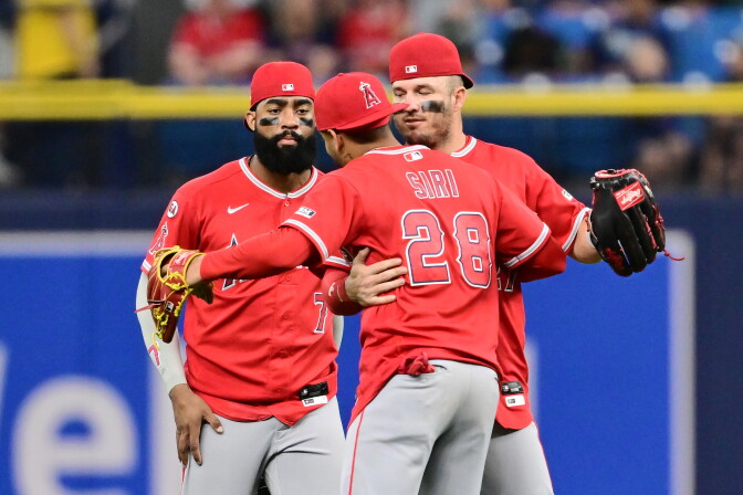 A Black man and a light-skinned man wearing red baseball uniforms hug a man with his back to the camera, also wearing a read baseball uniform with the number 28 and the name "Siri" on the back.