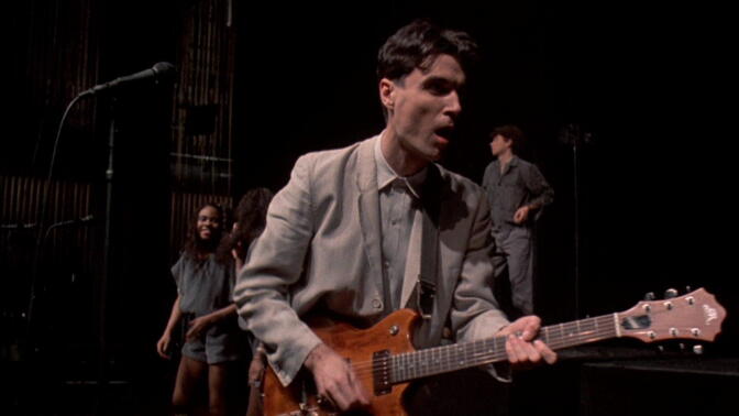 A light-skinned man in a gray suit plays electric guitar.