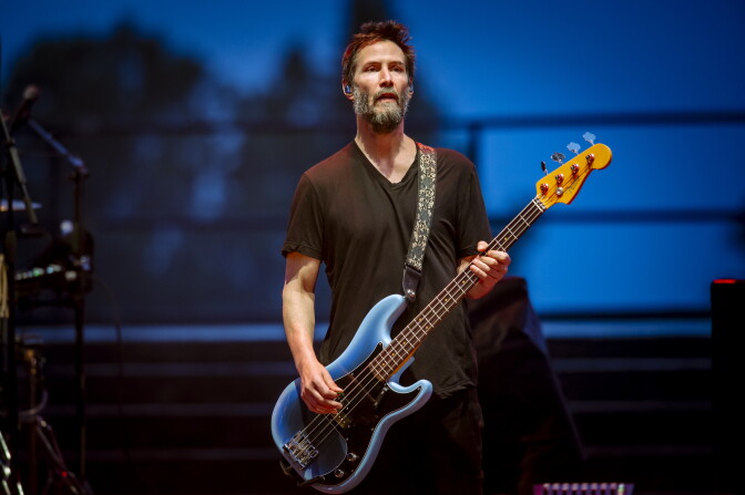 A light-skinned man with a beard plays a blue bass guitar onstage.