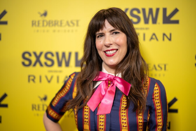 A light-skinned woman wearing a blue and gold striped shirt and a pink bow around her neck smiles at the camera.