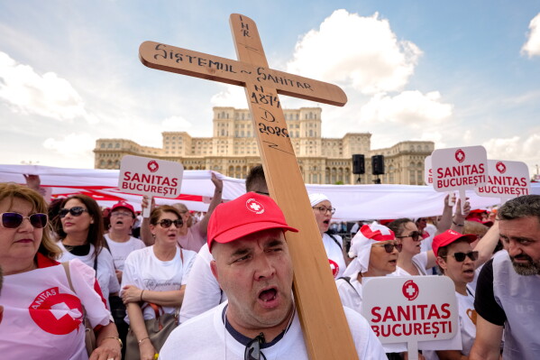 A healthcare worker holds a large cross reading "Medical system born 1874 deceased 2026" outside the Romanian Parliament, during a protest against planned changes into the legislation concerning public sector salaries, in Bucharest, Romania, Wednesday, June 3, 2026. (AP Photo/Vadim Ghirda)