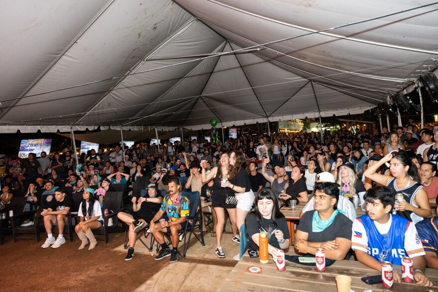 Hundreds of people in Spurs gear cheer underneath a large tent outdoors during the watch party.
