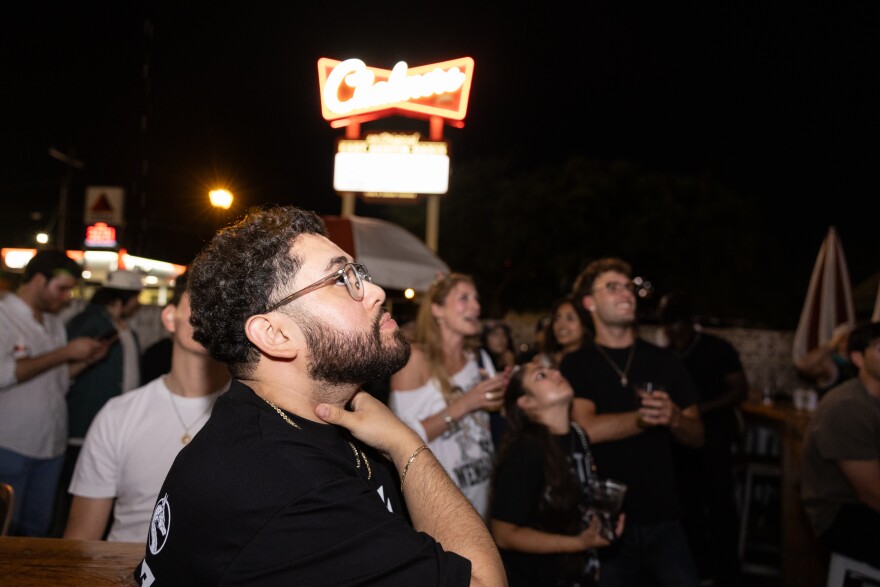A man with a concerned expression looks up at a TV screen at the watch party.