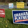 Campaign signs are staked in a grassy area with yellow flowers next to a road in Rockport, Maine.