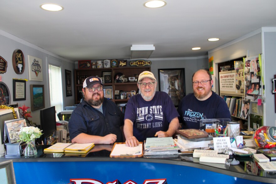 Three men stand behind the cluttered counter of a sign shop.