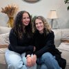 A teenager with curly, brown hair sits next to her mother who has shoulder-length, curly blonde hair, on a couch. They are both wearing black sweaters with blue jeans.
