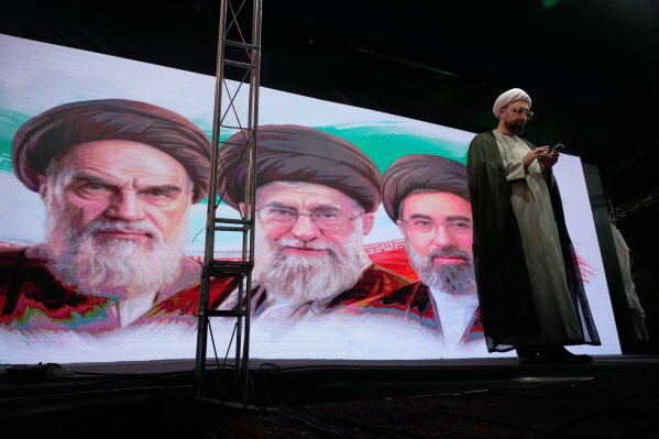 A cleric checks his cell phone on stage in front of a screen displaying portraits of the late Iranian revolutionary founder Ayatollah Khomeini, left, late Supreme Leader Ayatollah Ali Khamenei, center, and Supreme Leader Ayatollah Mojtaba Khamenei, during a pro-government gathering in Tehran, Iran, Sunday, June 7, 2026. (AP Photo/Vahid Salemi)