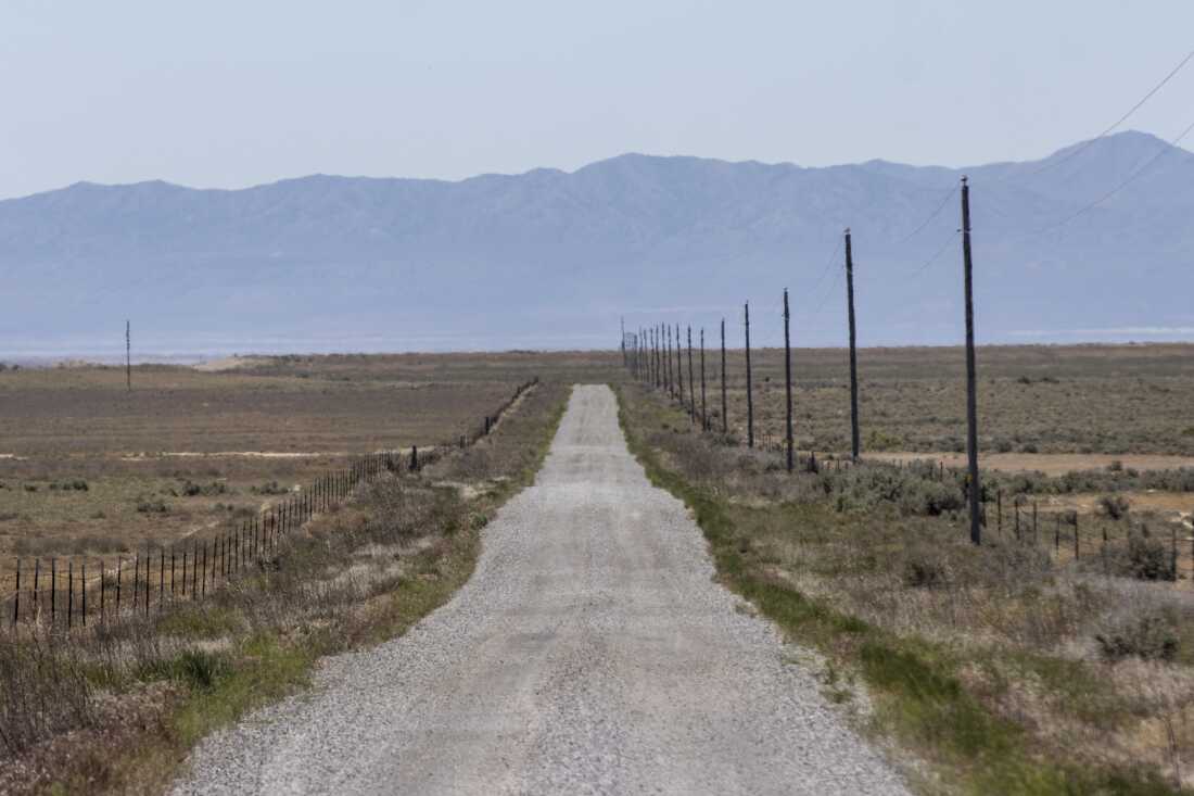 A gravel road stretches through the area where the Stratos Project, a proposed data center, will be built in Box Elder County near Snowville, Utah. There are mountains in the background. There is a fence running on the left of the empty gravel road, and power lines and fence running on the right.