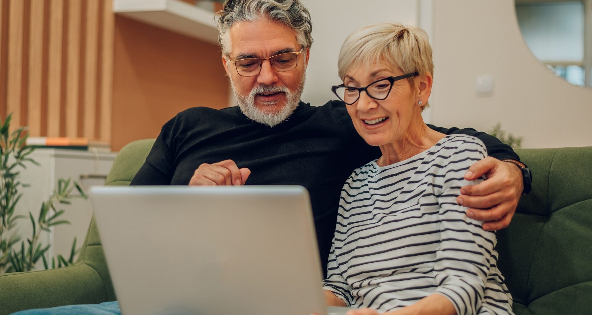Senior couple leaning towards each other using laptop while sitting on a couch in their home stock photo