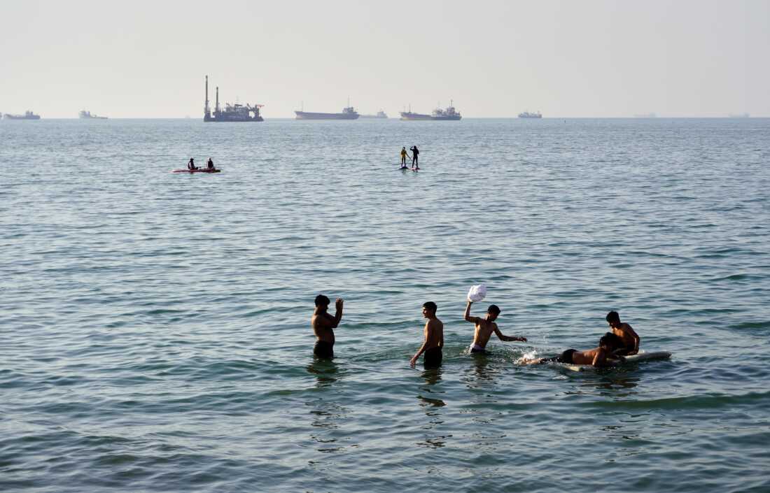 Residents swim and play in the waters of the Strait of Hormuz while cargo ships and commercial vessels lie anchored in the distance off Bandar Abbas, Iran, Wednesday, June 10, 2026.