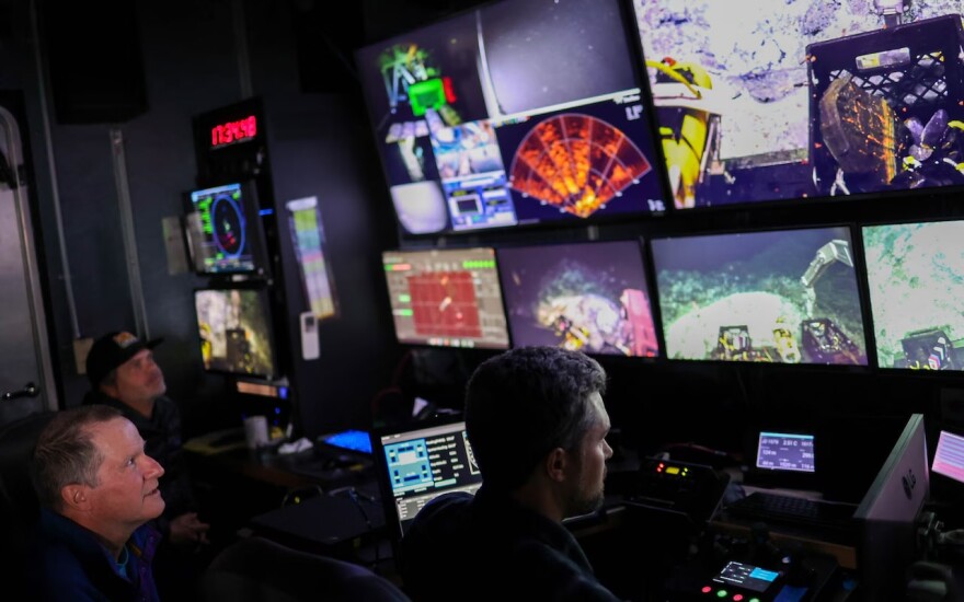 Three men look at a rack of video monitors in a dark room.