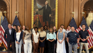 A group of students and staff pose for a photo inside a formal government building beneath a large portrait of George Washington. American flags flank the group as they stand together in an ornate room with wood-paneled walls and decorative furnishings.