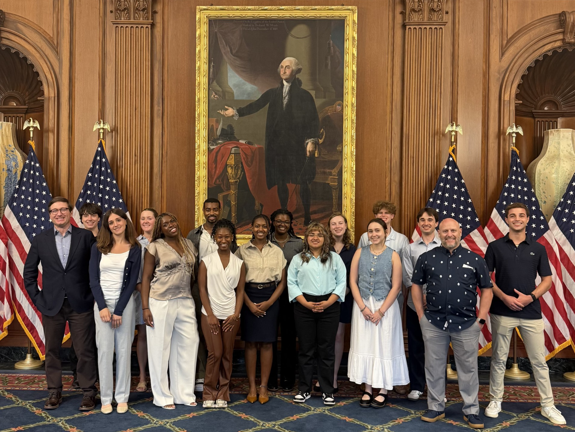A group of students and staff pose for a photo inside a formal government building beneath a large portrait of George Washington. American flags flank the group as they stand together in an ornate room with wood-paneled walls and decorative furnishings.