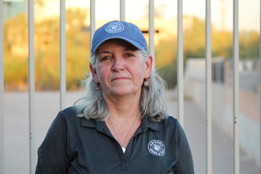 A woman wearing an Arizona Game & Fish polo shirt and baseball cap standing outside in front of a wrought iron fence