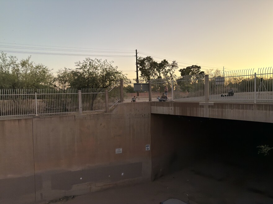 Three people sit and one stands behind a wrought-iron fence above the entrance to a large concrete flood-control tunnel