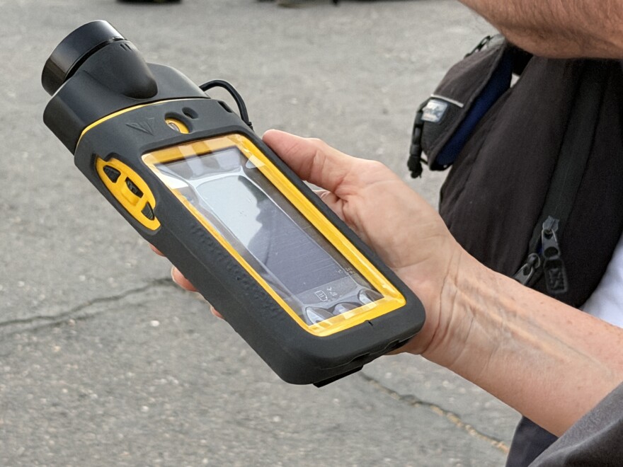A hand holding a medium-size gray and yellow device with an lcd screen, a rugged case and a specialized microphone attached to the top