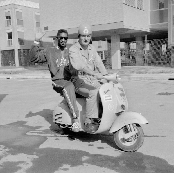 U.S. high jumper John Thomas, of Cambridge, Mass., waves from a Vespa scooter with an unidentified official Olympic driver inside the Olympic Village, in Rome, Aug. 27, 1960. (AP Photo/Mike Stern, File)