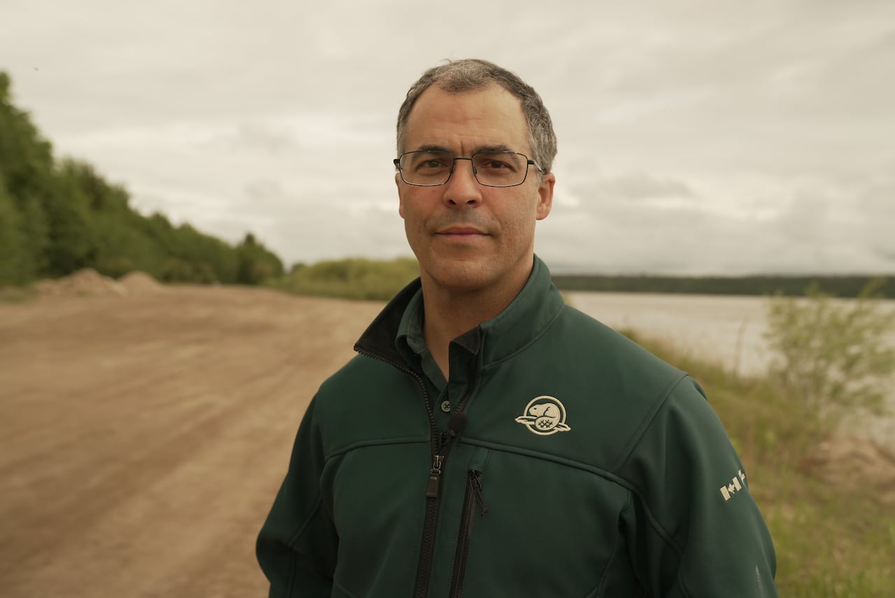A man in glasses and a green coat with a collar stares head-on at the camera while a dirt road is seen in background