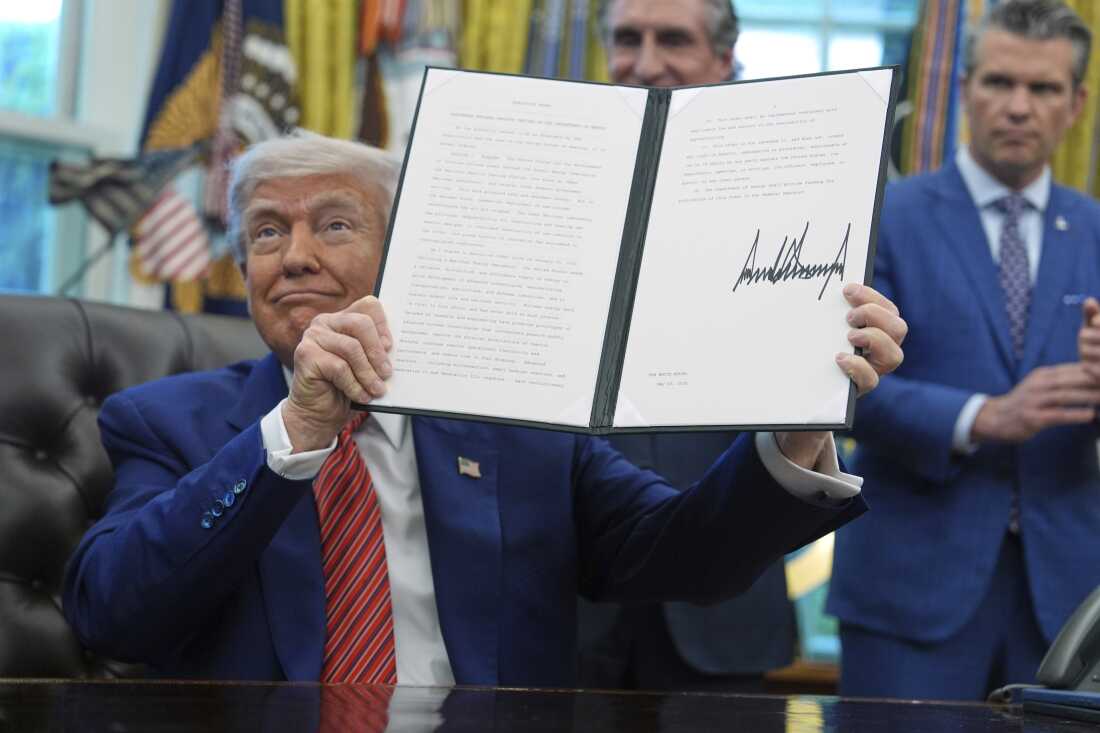 In this photo, President Trump holds up an executive order he signed. He's seated at a desk in the Oval Office of the White House on May 23, 2025.