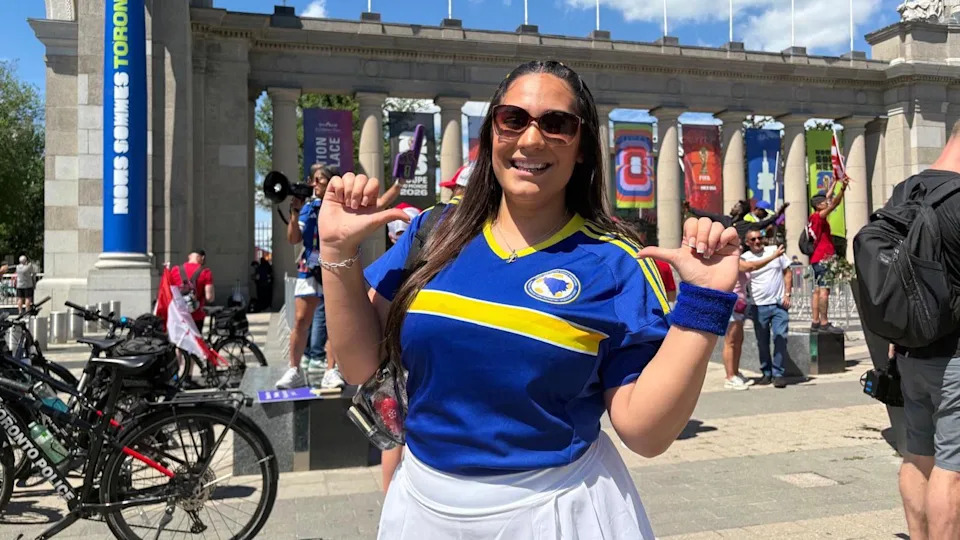 Layla is pictured wearing a Bosnian jersey and a white skirt with sunglasses. She has long black straight hair. Behind her are the gates to Toronto's stadium grounds, decorated with Fifa 2026 banners.