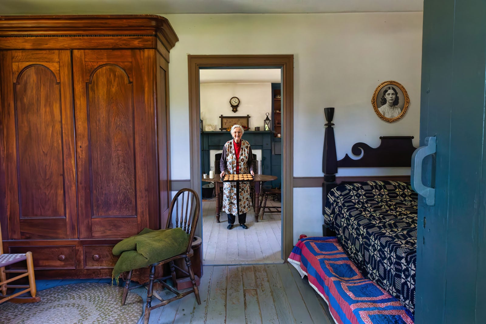An elderly person in period clothing stands in a doorway holding a tray, inside a vintage room with wooden furniture, a bed with a colorful quilt, and a framed portrait on the wall.