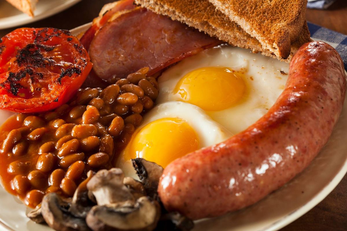 A white plate displays a traditional breakfast featuring a combination of fried eggs, sausages, baked beans, sliced tomatoes, and grilled mushrooms. The plate rests on a wooden surface.