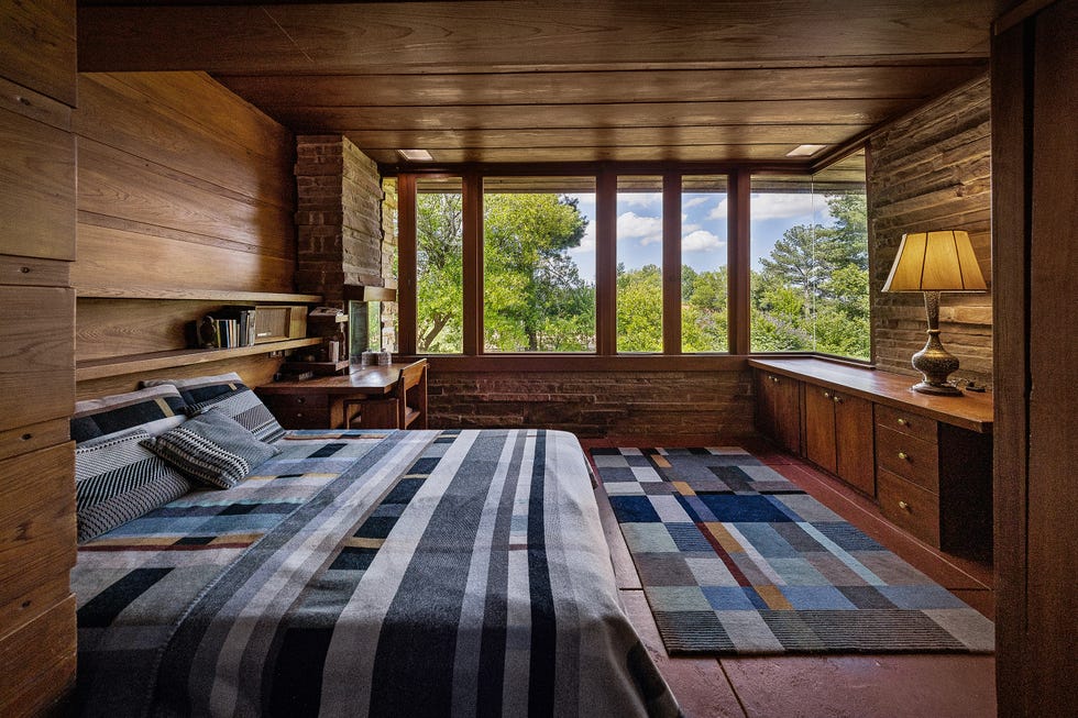 Cozy bedroom with wooden walls, large windows, a patterned blanket, and a stylish lamp.