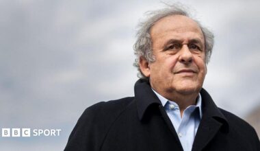 Michel Platini, silver-haired and wearing a black coat over a pale blue shirt, looks on