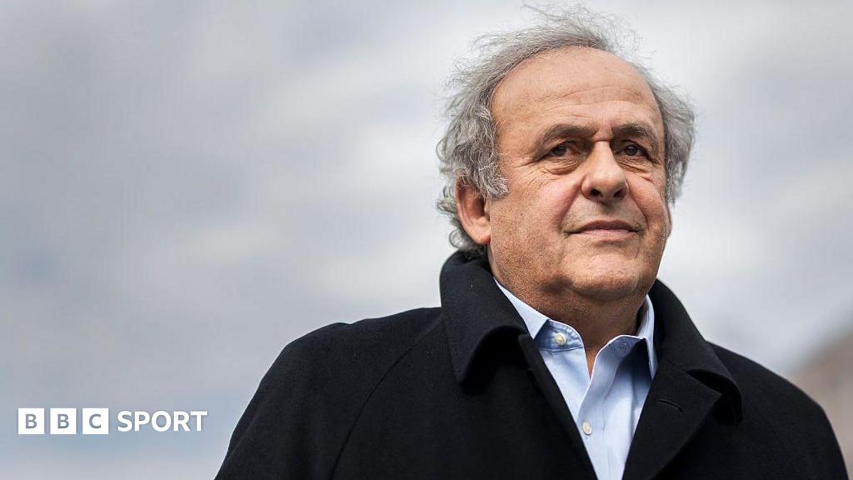 Michel Platini, silver-haired and wearing a black coat over a pale blue shirt, looks on