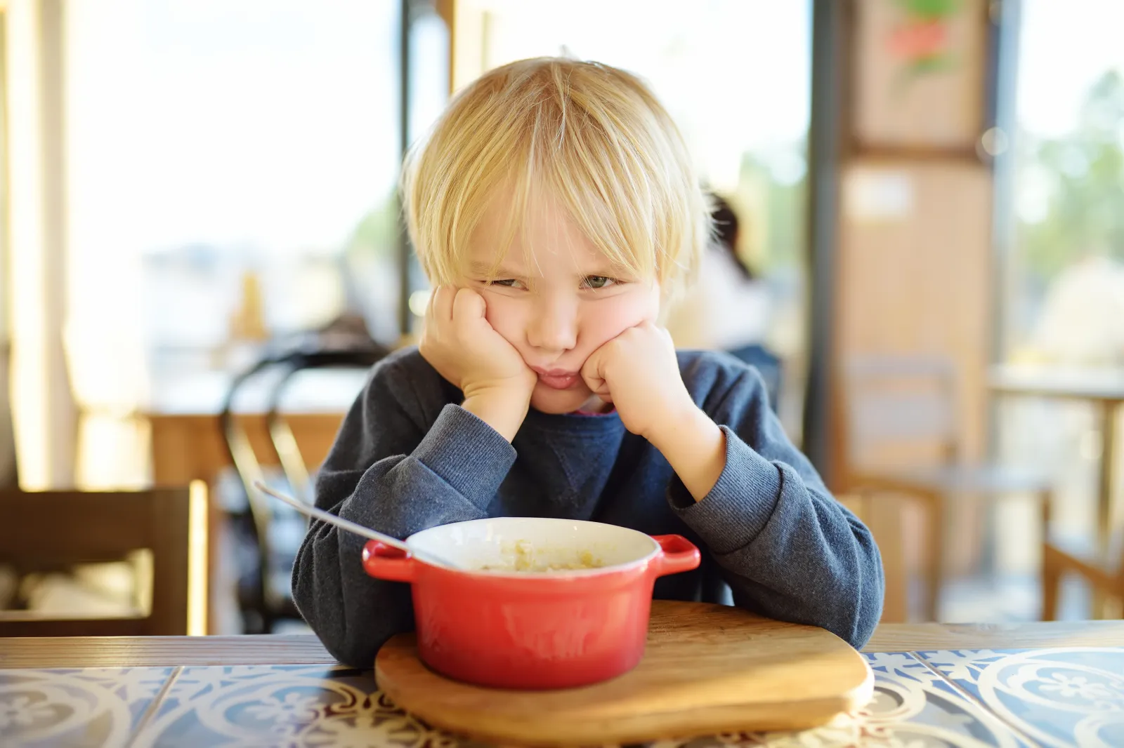 A stock photo of a young boy sitting at the table in cafe or restaurant and doesn't want to eat.