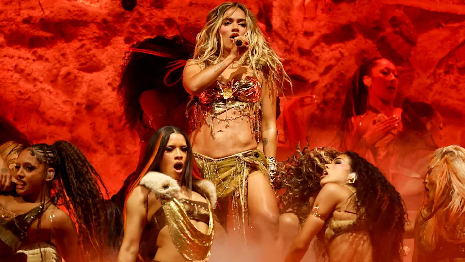 Karol G performs on stage in a dynamic outfit with dancers, set against a fiery red background.