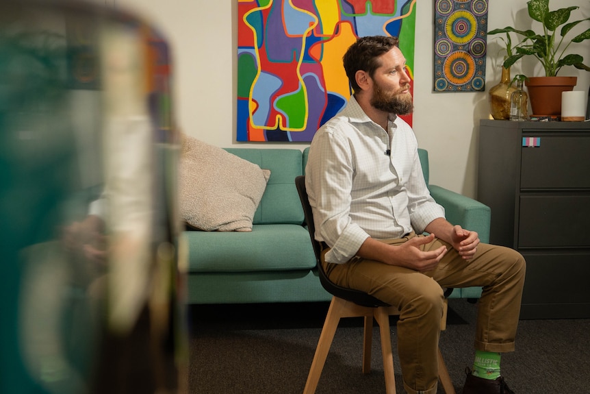Wide-shot image of white man, brown beard and hair, sitting on chair (right of frame), mid talking, hands in motion on lap.