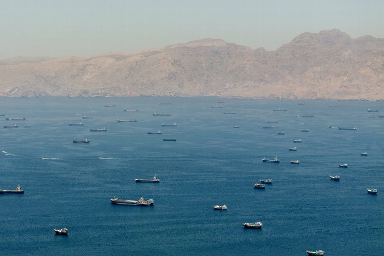 An aerial view of dozens of large ships anchored in a waterway in front of tan mountains.