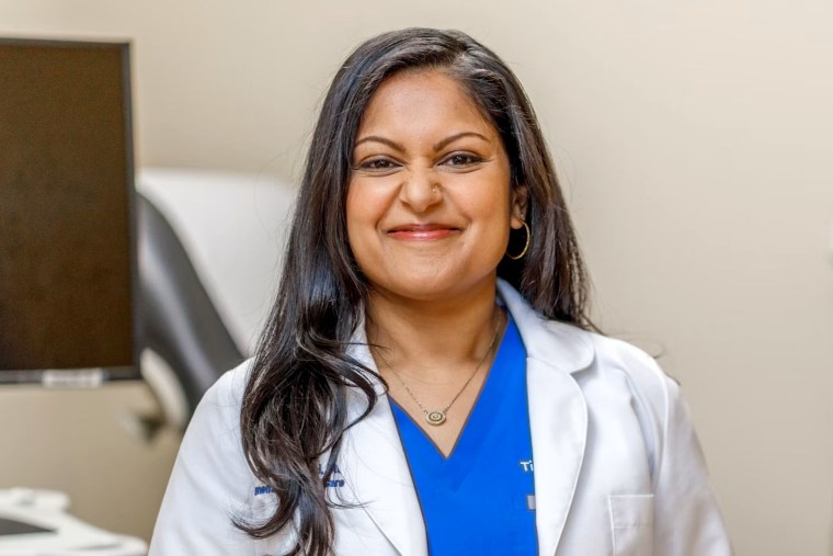 Tina Shah in a doctors white coat and scrubs.