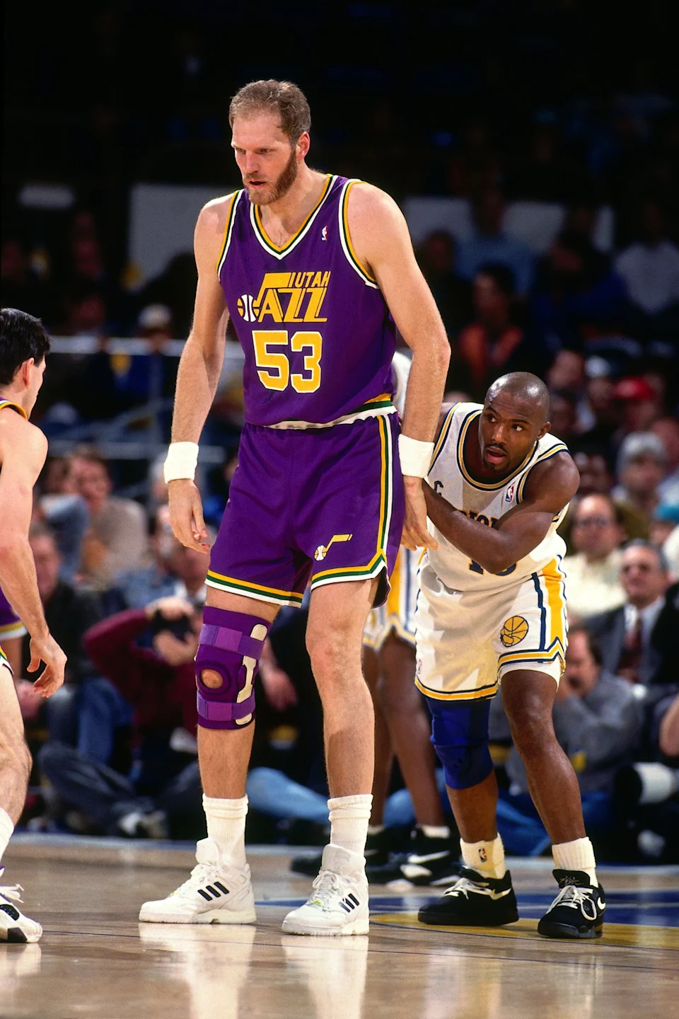 Basketball game action; a tall player in a Jazz uniform with number 53 wears a knee brace, guarded closely by a player in a Warriors uniform