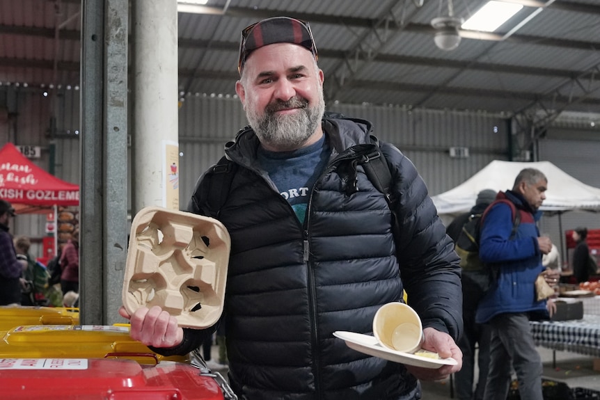 A man wearing a cap holding up compostable packaging in a warehouse surrounded by market stalls.